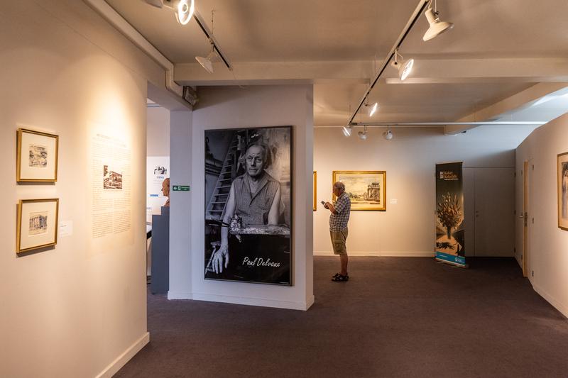 Interieur van een kunstgalerij met een zwart-wit portret dat prominent aan de muur hangt. Een man bekijkt in de buurt kunstwerken, wat een contemplatieve sfeer creëert.