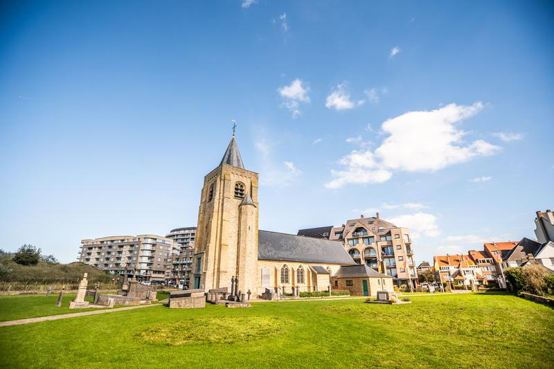 Historisch stenen kerkje met een hoge klokkentoren staat in een grasrijke omgeving onder een heldere blauwe lucht. Moderne gebouwen zijn op de achtergrond zichtbaar.