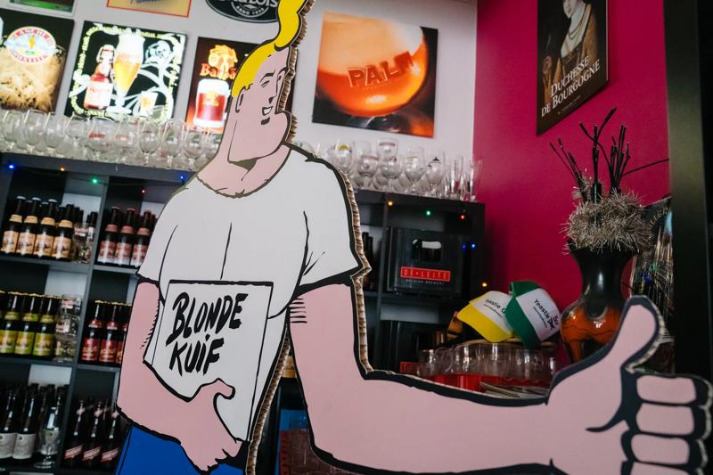 Pappaufsteller eines blonden Mannes mit Bier "Blonde Kuif" in einer Bar, mit Flaschen und Gläsern im Hintergrund.