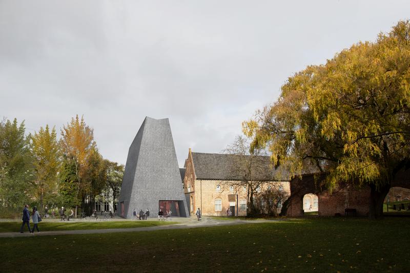 Modern gray building with a slanted, angular design beside historic brick structures, surrounded by autumn trees. People walk on a sunlit path.