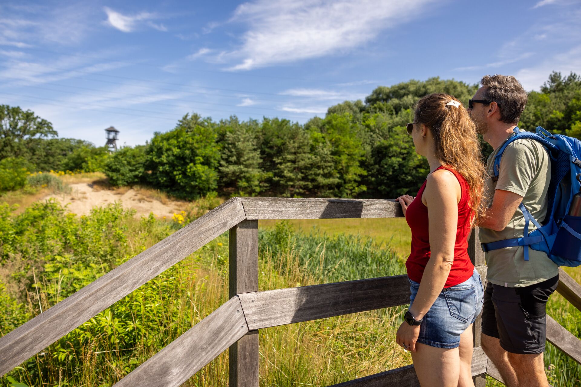 De schans van Waterschei Uomo e donna con zaini guardano da una piattaforma panoramica in legno su dune e bosco sotto un cielo blu.