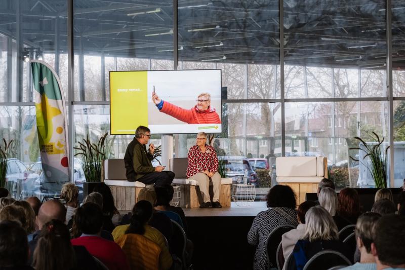 foto achteraan zaal genomen richting podium waarop 2 mensen zitten