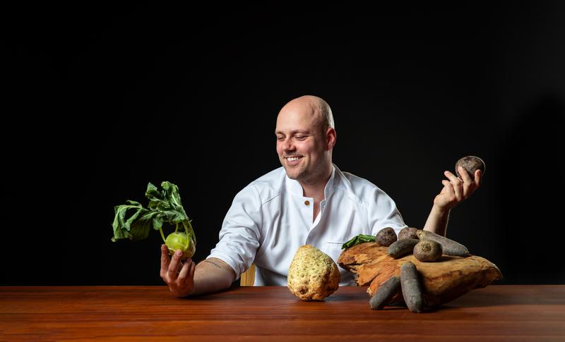 Een glimlachende chef in een witte jas houdt een koolrabi en een aardappel vast, zittend aan een houten tafel met diverse wortelgroenten, tegen een donkere achtergrond.