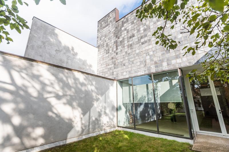 Exterior de un edificio moderno blanco con grandes ventanas y fachada de piedra geométrica, rodeado de árboles que proyectan sombras; una atmósfera tranquila y serena.