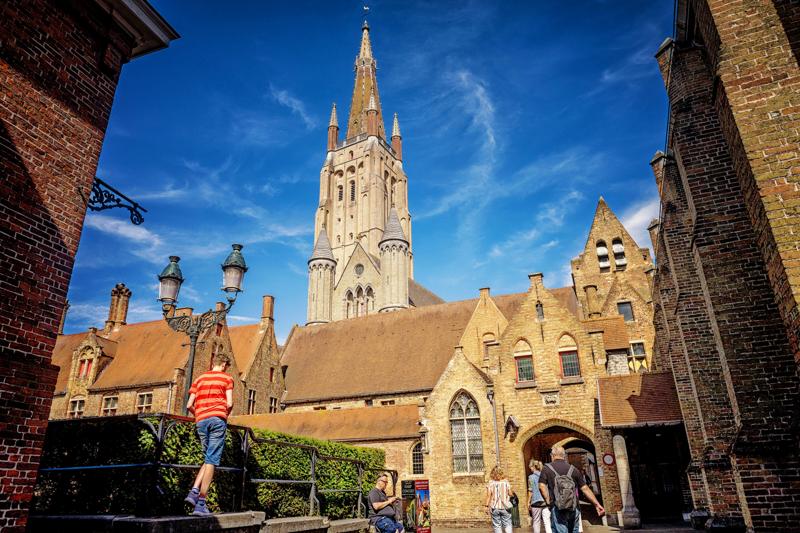 Medieval European square with a towering church spire under a clear blue sky. People walk on cobblestones, one child in a red-striped shirt by a hedge.