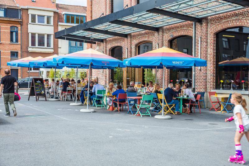 Buitencafé met kleurrijke stoelen en blauwe parasols op een zonnige dag. Mensen eten buiten, wat een levendige sfeer creëert. Een kind rolschaatst in de buurt.