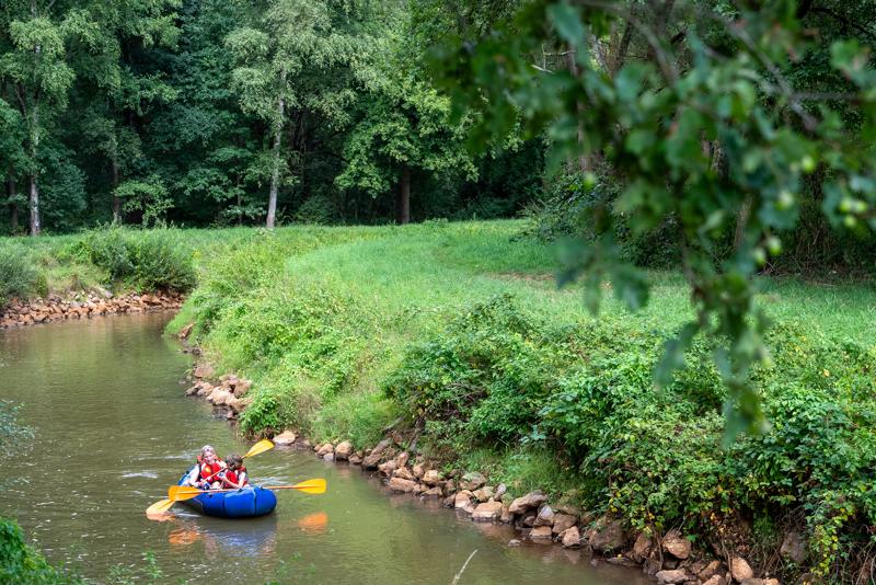 Deux personnes pagaient dans un bateau gonflable sur une rivière étroite le long d’une berge verte et boisée.