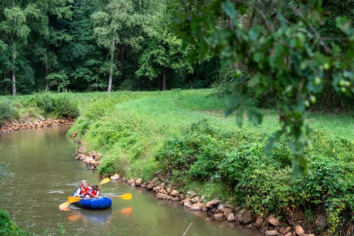 Bateau gonflable sur rivière en forêt Deux personnes pagaient dans un bateau gonflable sur une rivière étroite le long d’une berge verte et boisée.