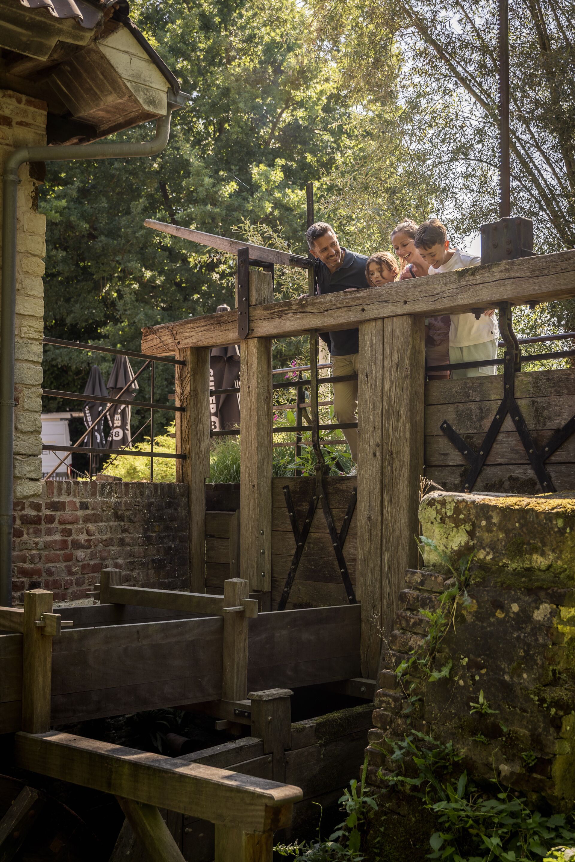 Een groep mensen leunt over een houten reling en bekijkt een oude watermolen omringd door weelderig groen. De scène straalt nieuwsgierigheid en rust uit.