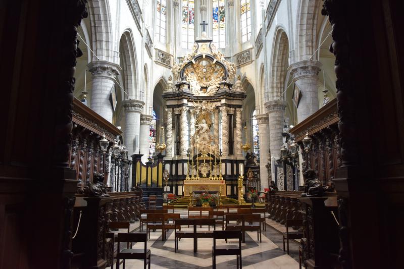 Intricate cathedral interior with ornate altar, towering columns, and carved wooden pews. Sunlight filters through stained glass windows, creating a serene atmosphere.