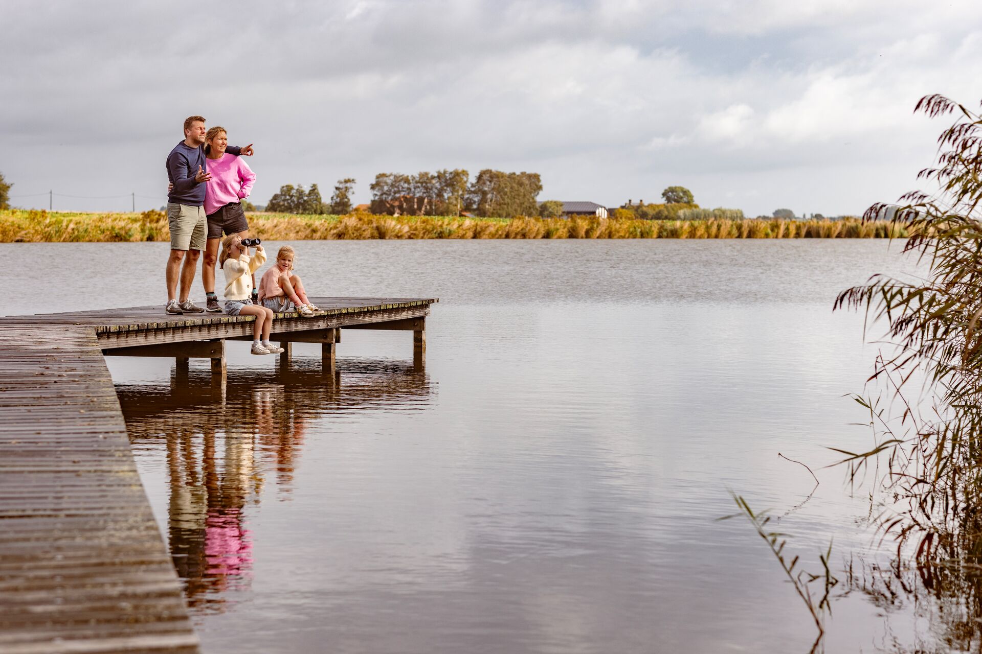 Een gezin met een hond geniet van het rustige uitzicht op een meer vanaf een houten steiger onder een bewolkte lucht. Een kind zit op de rand van de steiger, wat een serene, vredige sfeer creëert.