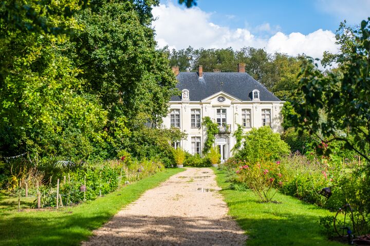 Statig wit landhuis met donker dak, genesteld tussen weelderige bomen, omgeven door levendige tuinen en een grindpad onder een helderblauwe lucht. Serene sfeer.