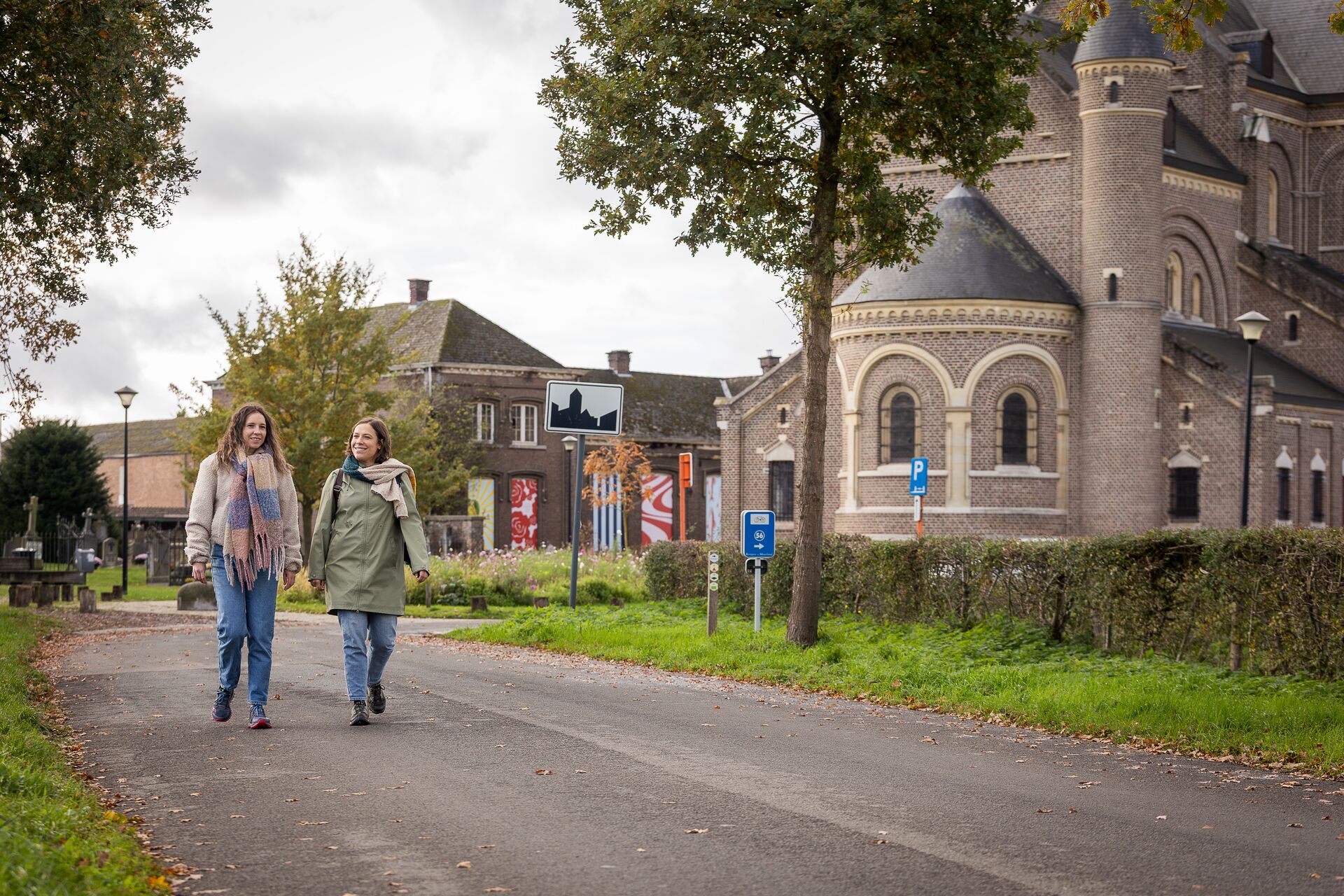 Dos mujeres caminan por una calle pavimentada junto a un edificio histórico de ladrillo con ventanas arqueadas. Es un día nublado y la escena transmite calma y tranquilidad.