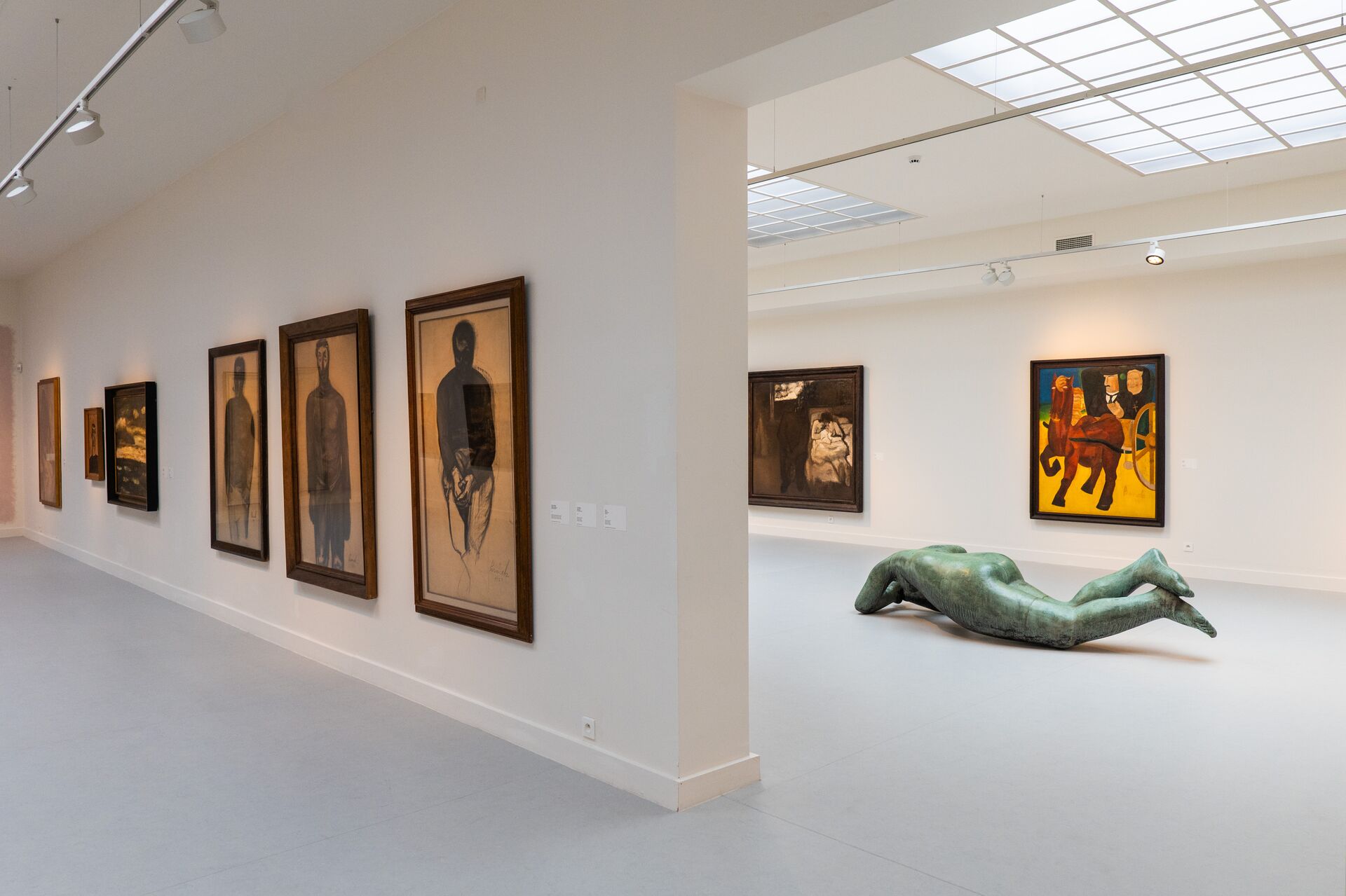 Una galleria d’arte moderna con dipinti astratti su pareti bianche. Una scultura umana verde e sdraiata giace sul pavimento, creando un’atmosfera serena.