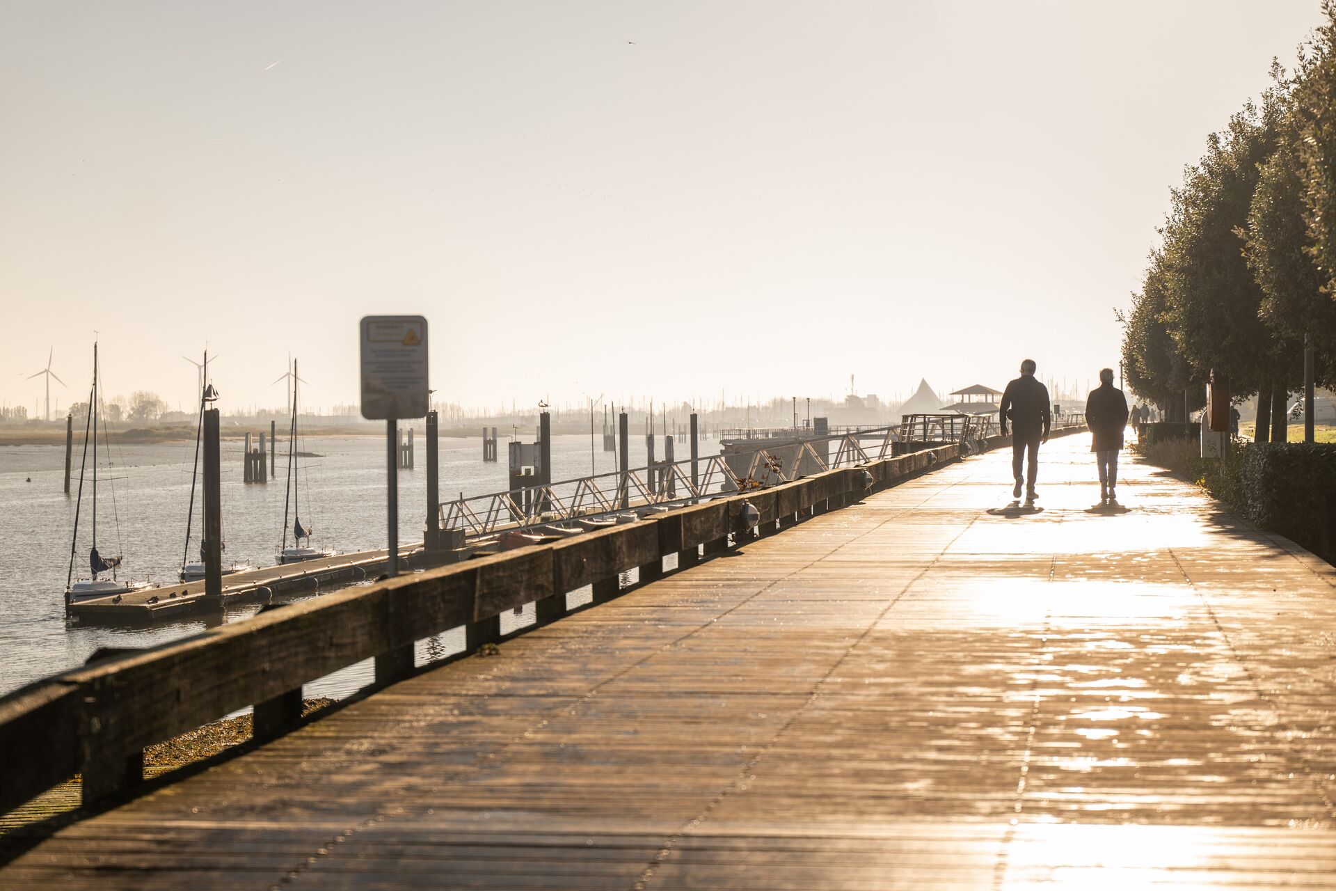 Vaargeul Nieuwpoort Een rustige wandelpromenade langs de rivier bij zonsopgang. Twee mensen wandelen over een zonovergoten pad met aan de ene kant bomen en aan de andere kant aangemeerde zeilboten op kalm water.