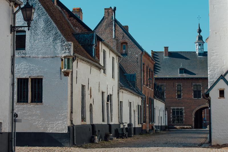 Historic cobblestone street lined with charming, old brick buildings, white facades, and arched windows, under a clear blue sky. Peaceful ambiance.