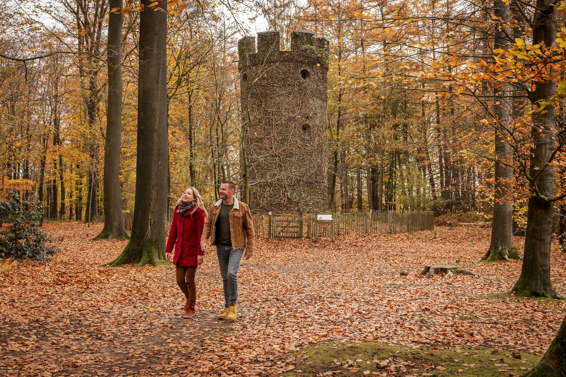 Una pareja camina por un bosque otoñal, con hojas esparcidas en el suelo. Detrás de ellos se alza una torre de piedra, rodeada de follaje colorido y dorado.