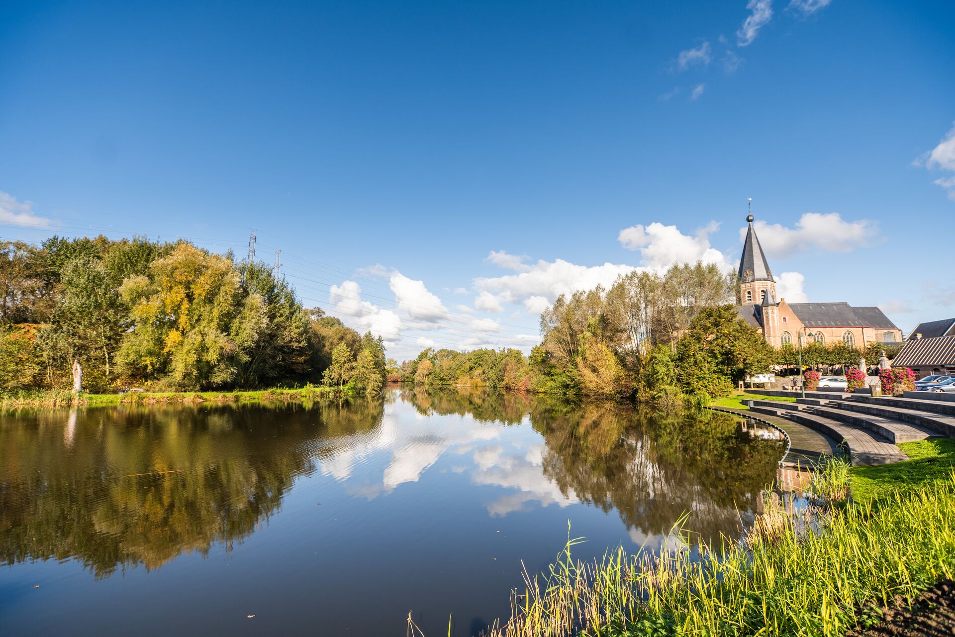A tranquil lakeside scene with a church and trees reflecting in the water under a clear blue sky. The peaceful setting conveys serenity and natural beauty.