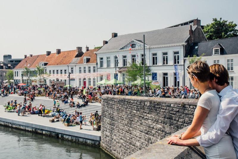 Een koppel kijkt uit over een levendige rivieroever. Mensen zitten op brede trappen, met kleurrijke historische gebouwen en een heldere lucht op de achtergrond, wat een vrolijke, zomerse sfeer oproept.