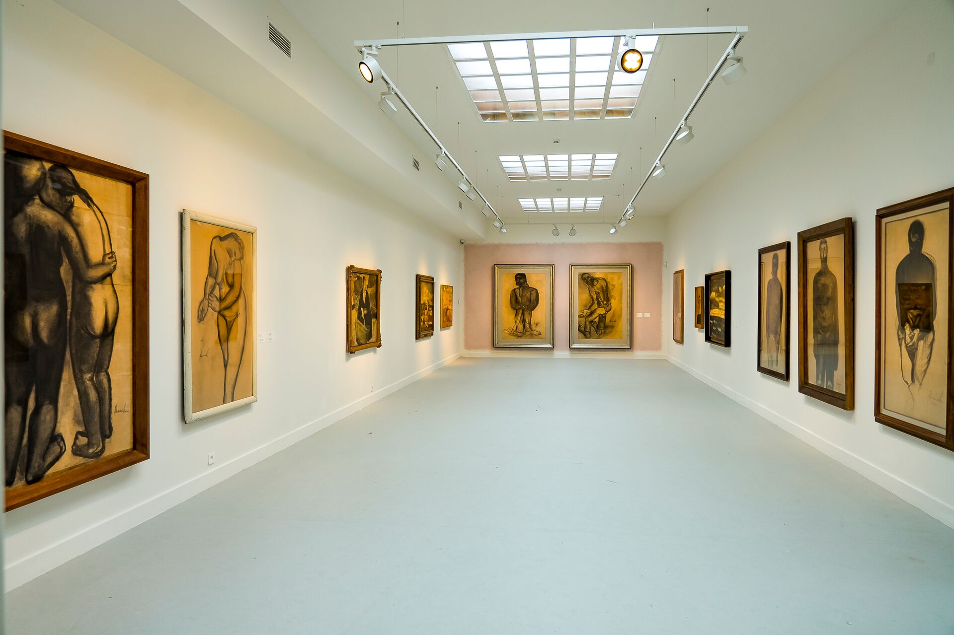 Long museum gallery with paintings and drawings on both walls, lit by skylights.