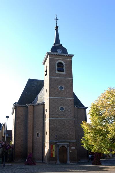 Een hoge bakstenen kerk met klokkentoren en klok onder een helderblauwe lucht. Zonovergoten boom rechts. Vredige en historische sfeer.