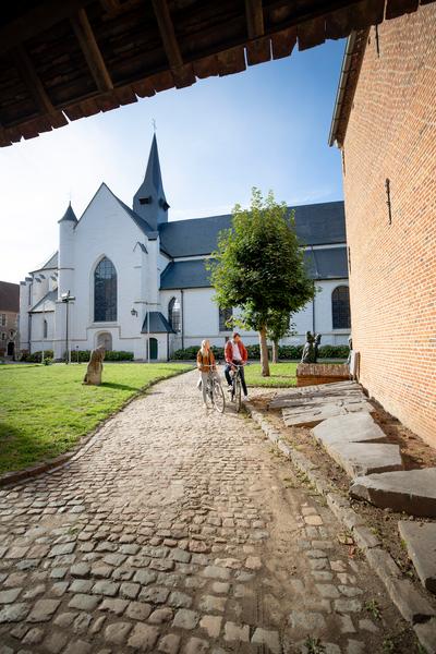 Een kasseienpad leidt naar een witte kerk met een hoge toren onder een strakblauwe lucht. Een groep mensen loopt ernaartoe, wat een serene sfeer oproept.