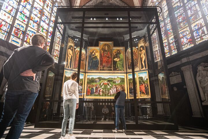 Bezoekers bewonderen een groot, gedetailleerd meerpanelen schilderij in een kathedraal, omringd door kleurrijke glas-in-loodramen, wat een plechtige sfeer creëert.