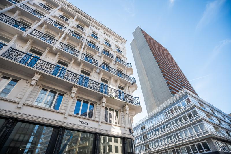 Escena urbana con un edificio clásico blanco con balcones azules ornamentados a la izquierda, contrastando con una torre moderna de vidrio y ladrillo a la derecha bajo un cielo despejado.