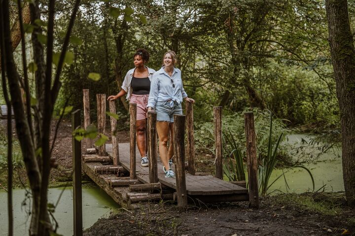Twee mensen lopen over een smalle houten brug boven een klein waterlichaam in een bosrijke omgeving. De omgeving is weelderig groen met bomen en struiken. De brug is gemaakt van houten planken en ondersteund door verticale houten palen.