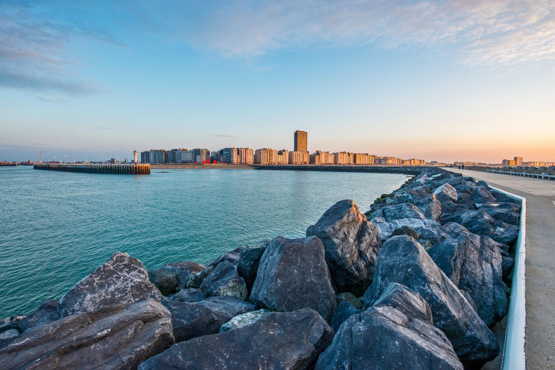Rock blocks on a pier with a calm sea and the skyline of Oostende at sunset.