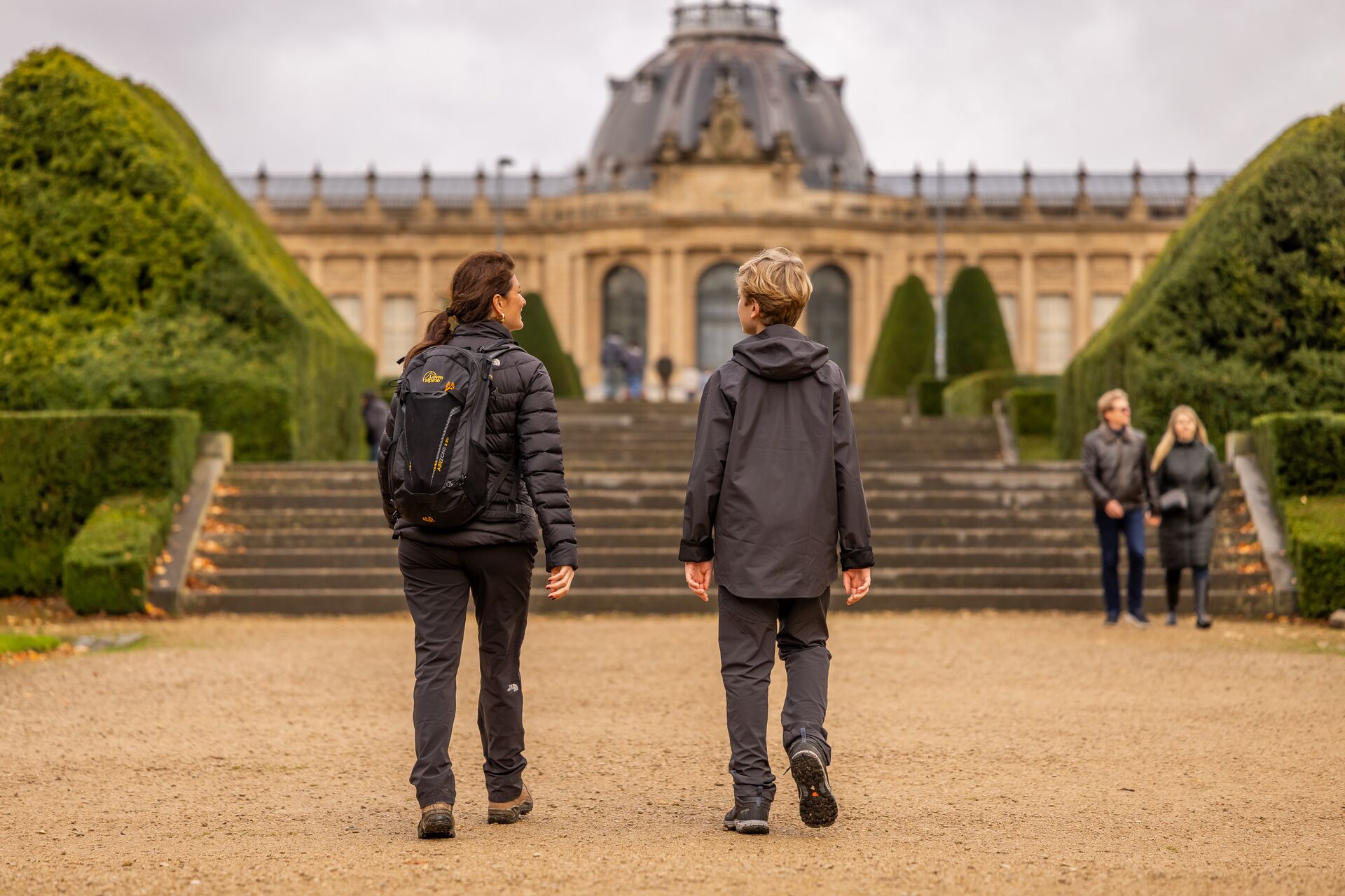 Due persone con giacche si dirigono verso un imponente edificio storico con cupola, circondato da siepi ben curate, sotto un cielo nuvoloso.