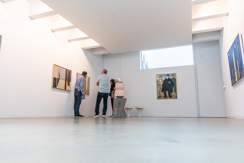 Drie mensen bewonderen abstracte kunst in een moderne, goed verlichte galerij, wat nieuwsgierigheid en waardering uitstraalt. Schilderijen en sculpturen sieren de minimalistische ruimte.