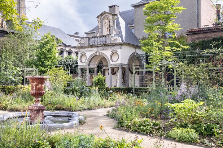 Rubenshuis Een historische tuin met weelderig groen omringt een rijk versierd gebouw met bogen en sculpturen. Een stenen pad leidt naar een centrale fontein, wat een serene sfeer oproept.