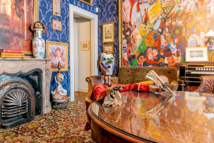 Eclectic living room with colorful mural, ornate fireplace, blue wallpaper, and mannequin in red robe at round table.
