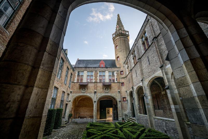 Historic courtyard framed by stone arch, featuring a medieval building with arched windows, a spire, and trimmed hedges. The sky is clear.