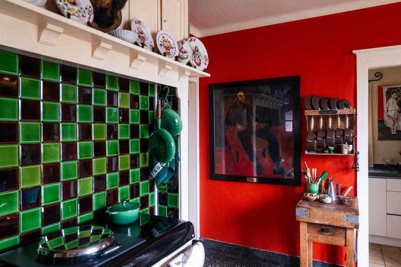 Una cucina vivace con pareti rosse sorprendenti presenta un paraschizzi di piastrelle verdi e nere, pentole appese, un bancone in legno rustico e piatti decorativi.  NL