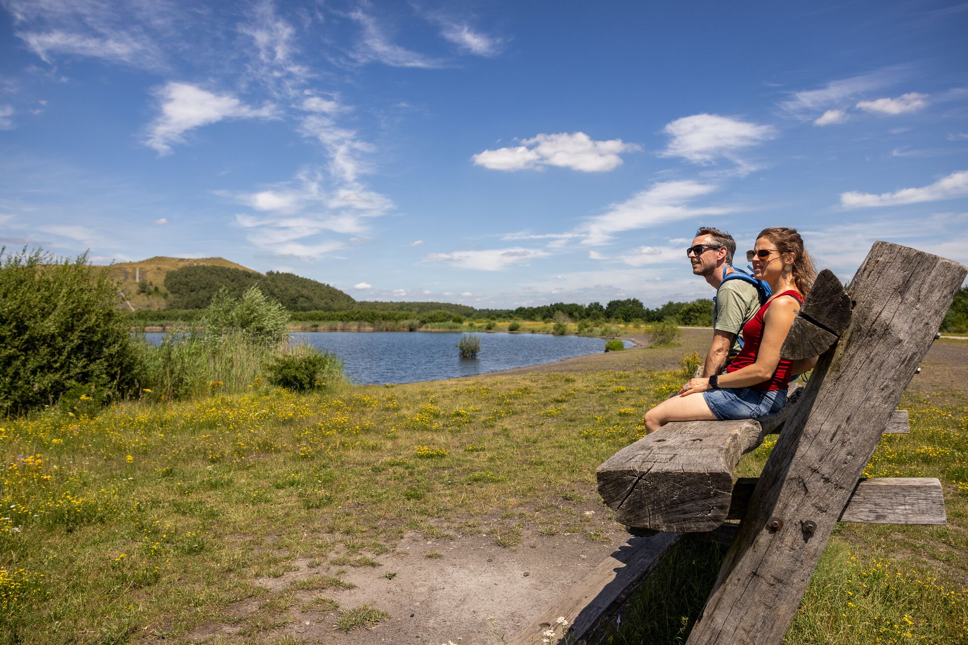 Escursionisti su panchina vicino a un lago Due escursionisti sono seduti su una panchina di legno e guardano un lago in un paesaggio verde sotto cielo azzurro.