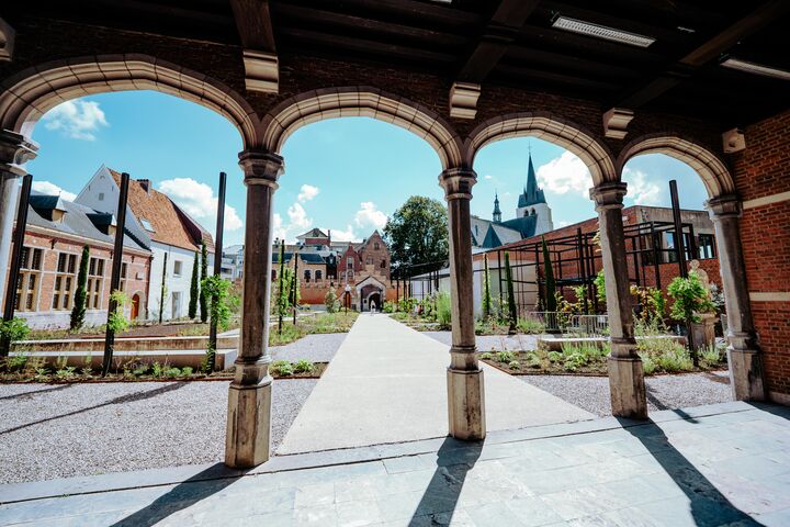 Binnenplaats Hof van Busleyden met bogen en kerktoren Binnenplaats met tuin en pad gezien door stenen bogen, met historische gebouwen en kerktoren op de achtergrond.