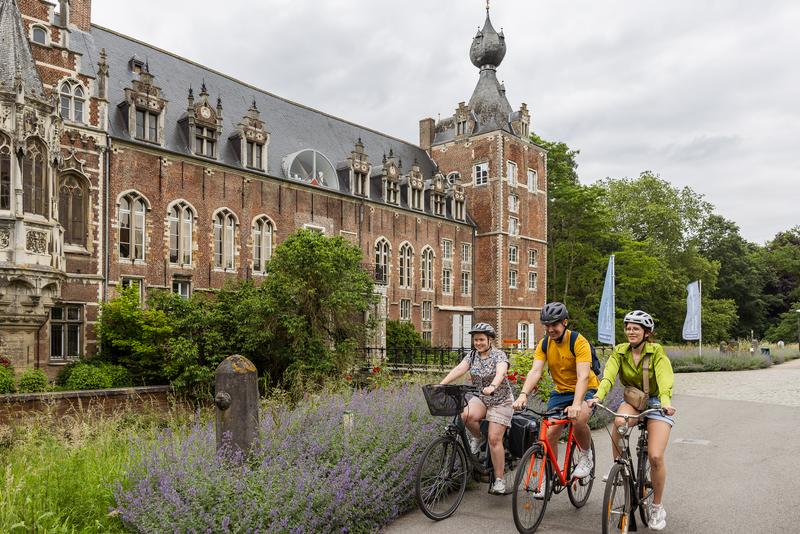 Three people ride bicycles past a historic building with ornate architecture and a tall tower. The scene is peaceful and surrounded by greenery.