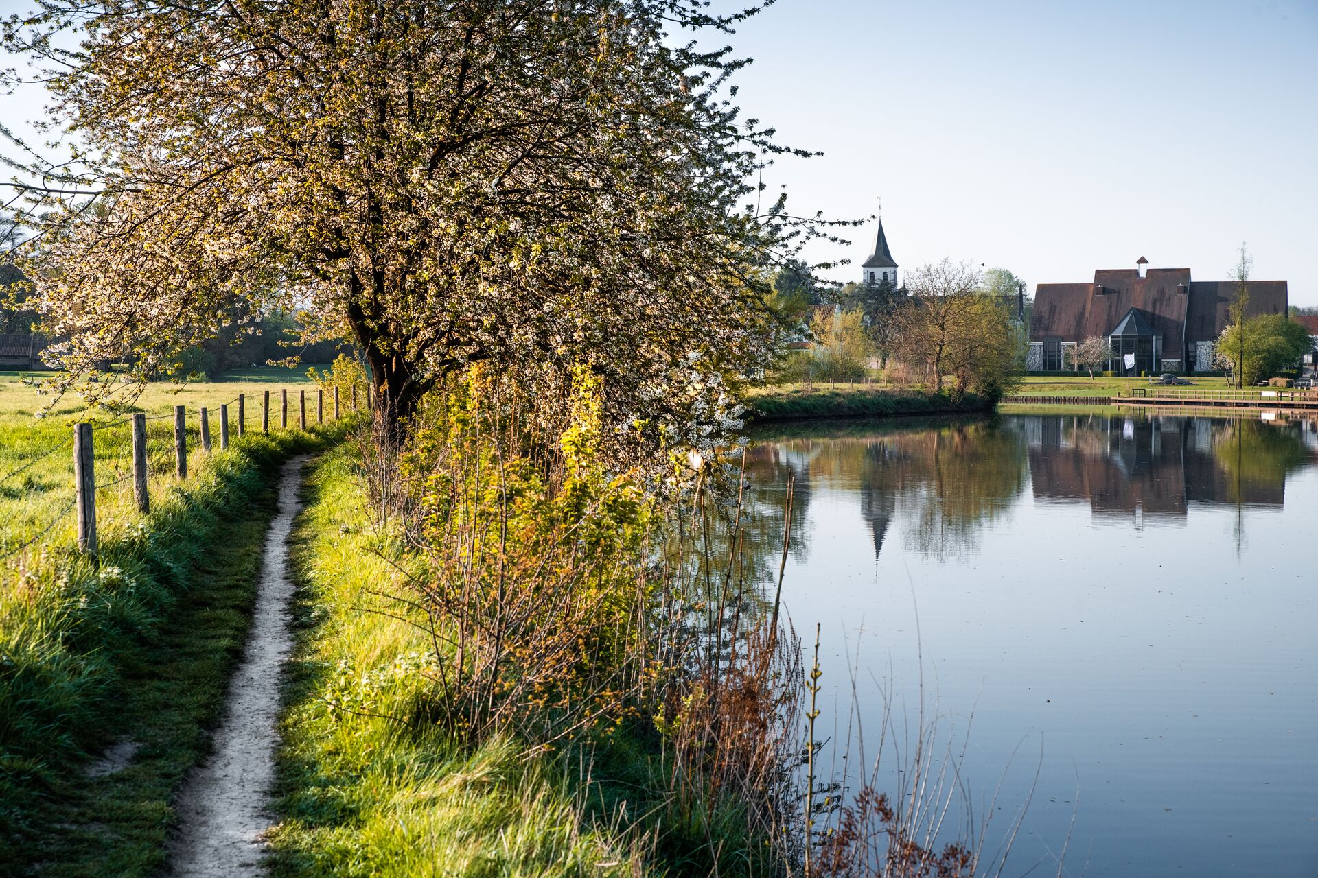 Sint-Martens-Latem Un paesaggio sereno con un sentiero stretto lungo un fiume tranquillo, fiancheggiato da alberi in fiore. In lontananza si vedono case pittoresche e un campanile. La scena evoca tranquillità e fascino rurale.