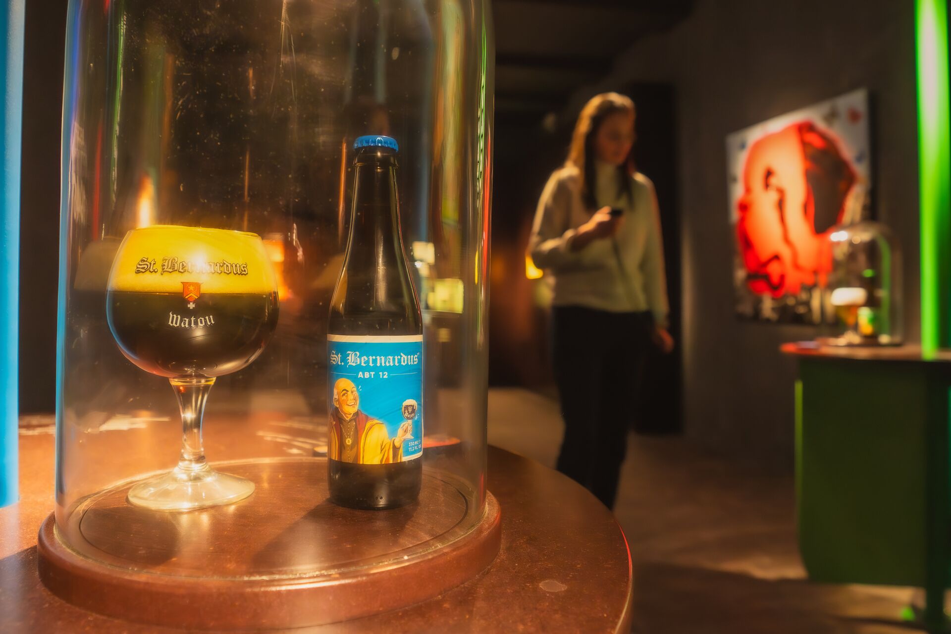 St. Bernardus beer glass under glass dome Glass dome with beer glass and St. Bernardus bottle on a pedestal; in the background a person in a museum room.