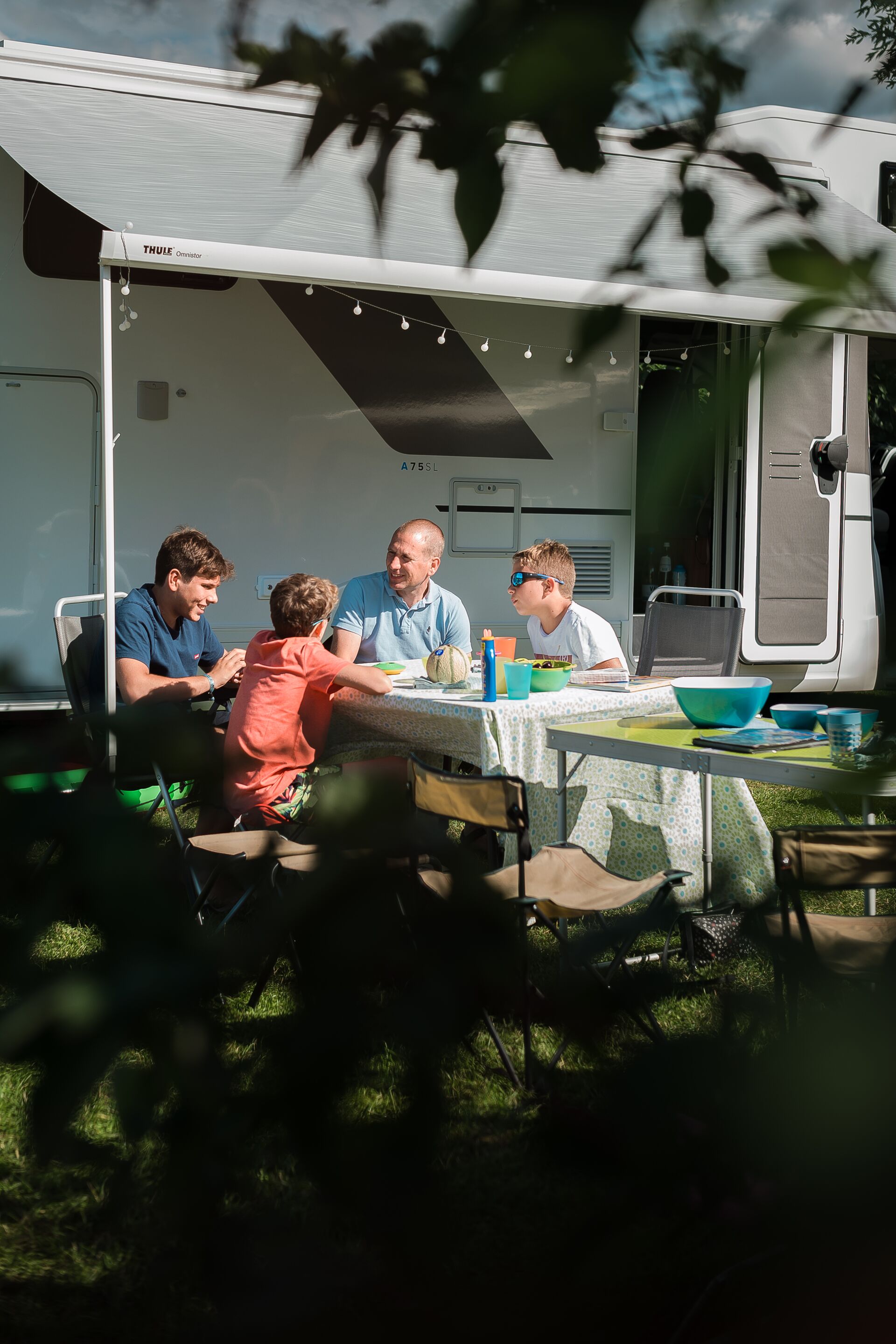 Een familie zit voor een camper te ontbijten aan een eettafel.