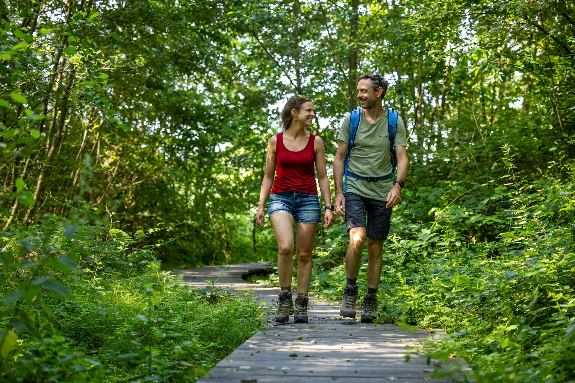 Escursionisti su passerella nel bosco Due escursionisti camminano su una passerella di legno in un bosco verde e si sorridono.