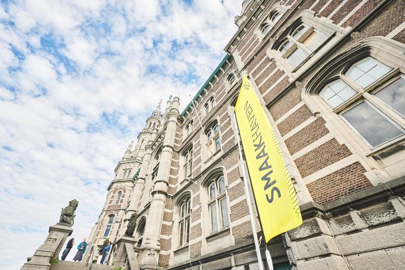 Een historisch, sierlijk gebouw met gebogen ramen en gedetailleerd metselwerk tegen een blauwe lucht. Voor het gebouw hangt een geel spandoek met tekst, wat een culturele setting weergeeft.