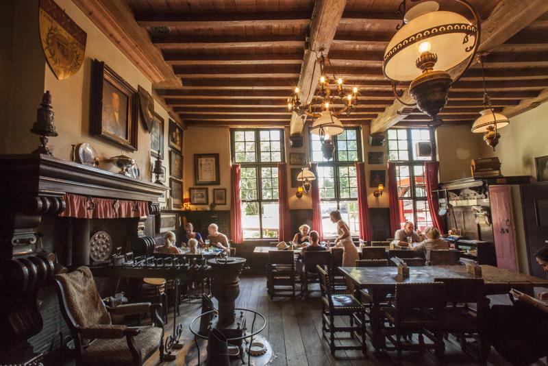 Cozy rustic tavern with wooden beams, vintage chandeliers, and red curtains. Patrons dine at wooden tables, creating a warm, inviting atmosphere.