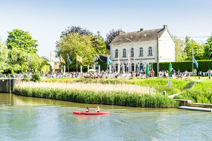 Astene - Deinze Un kayak rosso con due persone che remano su un fiume, con una riva erbosa e una grande casa bianca sullo sfondo. La scena è luminosa e vivace.