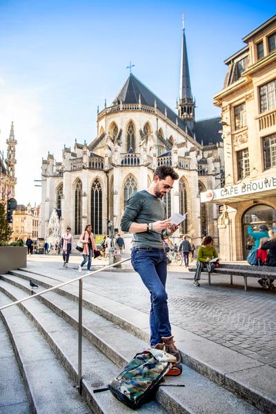 Een man staat op de trappen van een Europees plein een kaart te lezen, met een gotische kerk en mensen op de achtergrond. Een zonnige dag zorgt voor een levendige, ontdekkende sfeer.