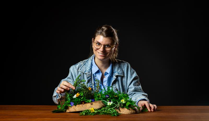 A person in a denim jacket and glasses smiles while arranging vibrant flowers on a wooden table against a black background, exuding creativity and joy.