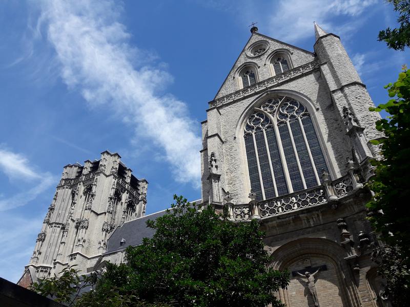 A towering Gothic cathedral façade and bell tower against a bright blue sky, adorned with ornate stone details and large arched windows, creating a majestic atmosphere.