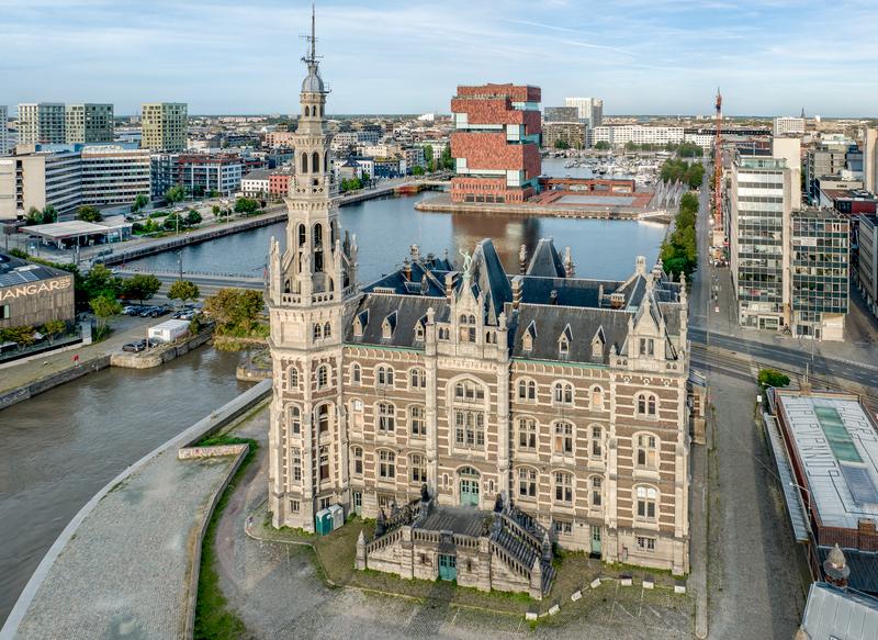 Bovenaanzicht van het Loodswezen in Antwerpen, een historisch gebouw met een hoge, sierlijke toren. Eromheen liggen moderne gebouwen, kanalen en een rood museum. De scène straalt een mix van geschiedenis en moderniteit uit.